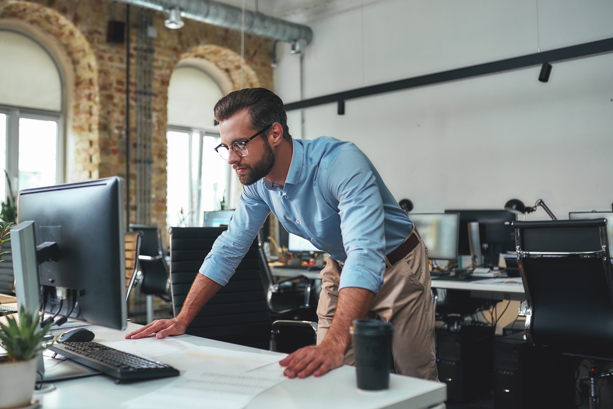 Busy day. Young bearded businessman in eyeglasses and formal wear using computer while standing in
